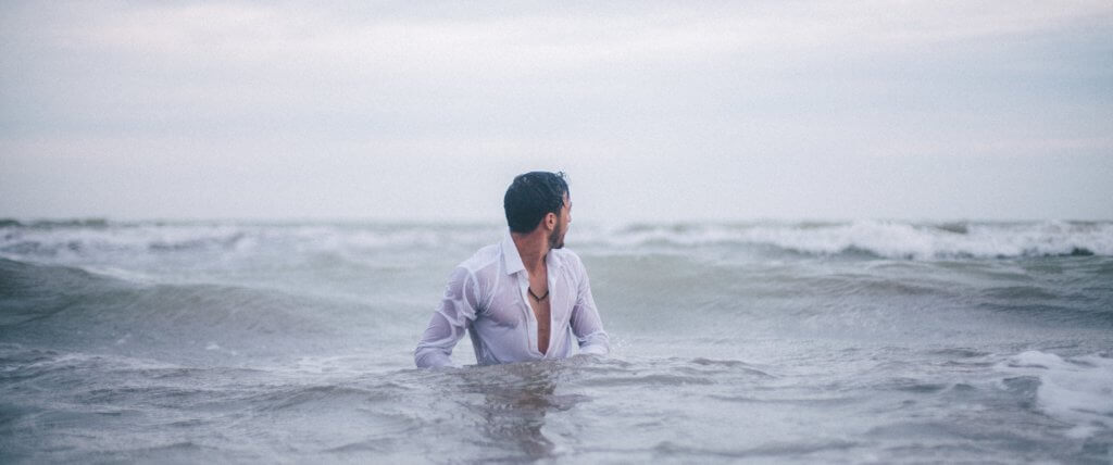 Bearded man with white shirt standing in rough water.